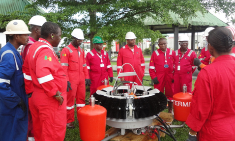 Workers in red coveralls gather around oil spill recovery equipment outdoors.