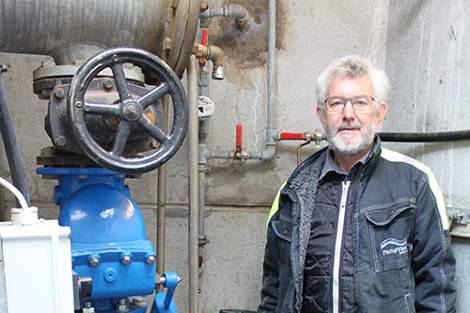Man standing beside industrial valve and piping in a mechanical setting.