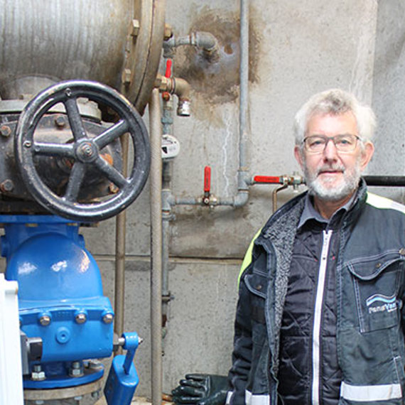 Man standing beside industrial valve and piping in a mechanical setting.