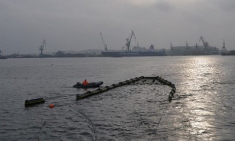 Boat near oil boom in harbor with cranes and ship in the background.