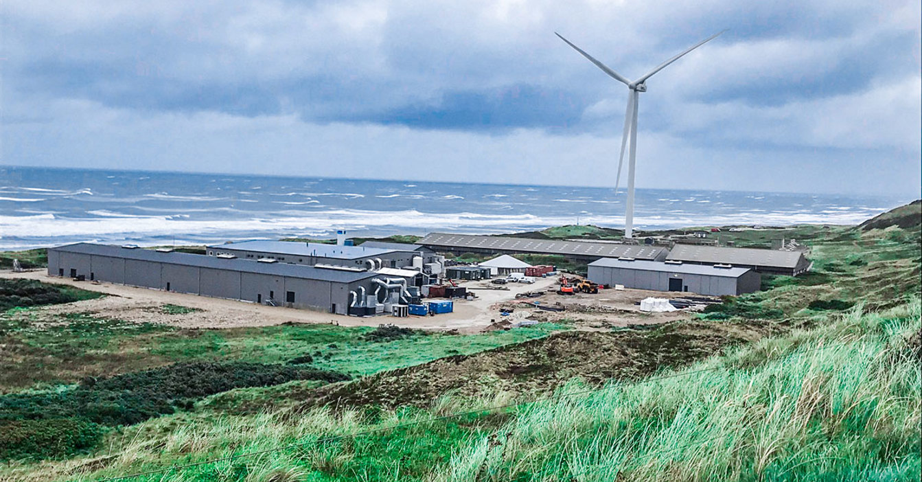 Wind turbine near industrial facility by the sea, surrounded by green landscape.