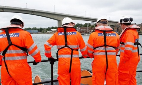 Four workers in orange DESMI RosClean suits on a boat near a bridge.