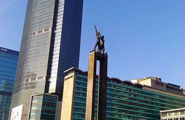 Tall skyscraper and monument with fountains in urban setting under clear blue sky.