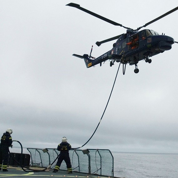Helicopter refueling on a ship's deck, with two crew members assisting.