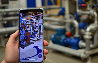 Hand holding smartphone showing augmented reality in industrial setting with pipes.