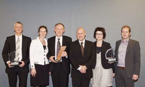 Six people posing with awards in formal attire against a plain background.