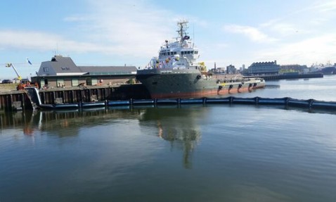 Ship docked at a harbor with floating barriers on a sunny day.