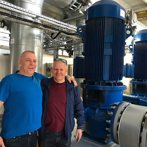 Dwóch mężczyzn stojących w przemysłowym otoczeniu z dużymi niebieskimi pompami.