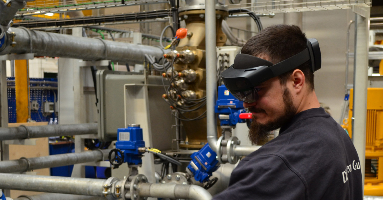 Technician wearing augmented reality headset in industrial facility with pipes and machinery.
