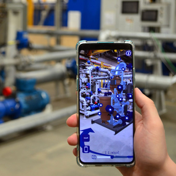 Hand holding smartphone with augmented reality view of industrial pipes and machinery.