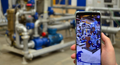 Hand holding smartphone with augmented reality view of industrial pipes and machinery.