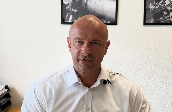 Man in white shirt speaking, with industrial photos on the wall behind him.