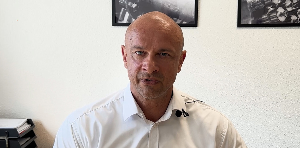 Man in white shirt speaking, with industrial photos on the wall behind him.