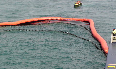 Orange oil containment boom deployed at sea with a small boat nearby.
