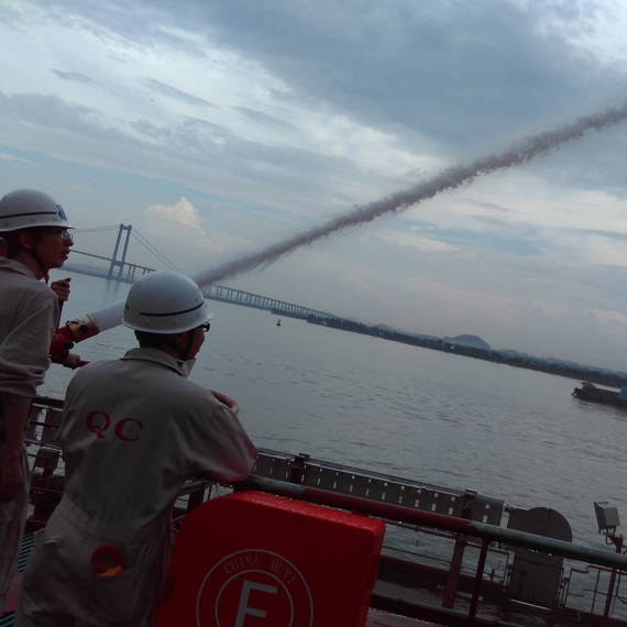 Personer använder en brandslang på en båt, med en bro i bakgrunden.