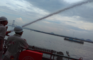 Personer använder en brandslang på en båt, med en bro i bakgrunden.