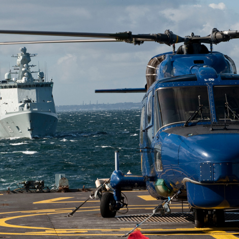 Blue helicopter on deck with a naval ship approaching in the background.