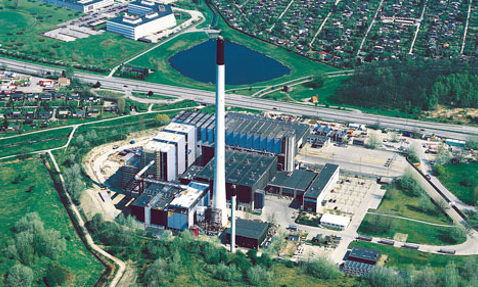 Flygfoto av en stor industrianläggning med en hög skorsten och omgivande grönska.