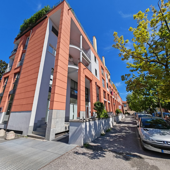 Modern orange building with large windows, cars parked nearby, and trees lining the street.