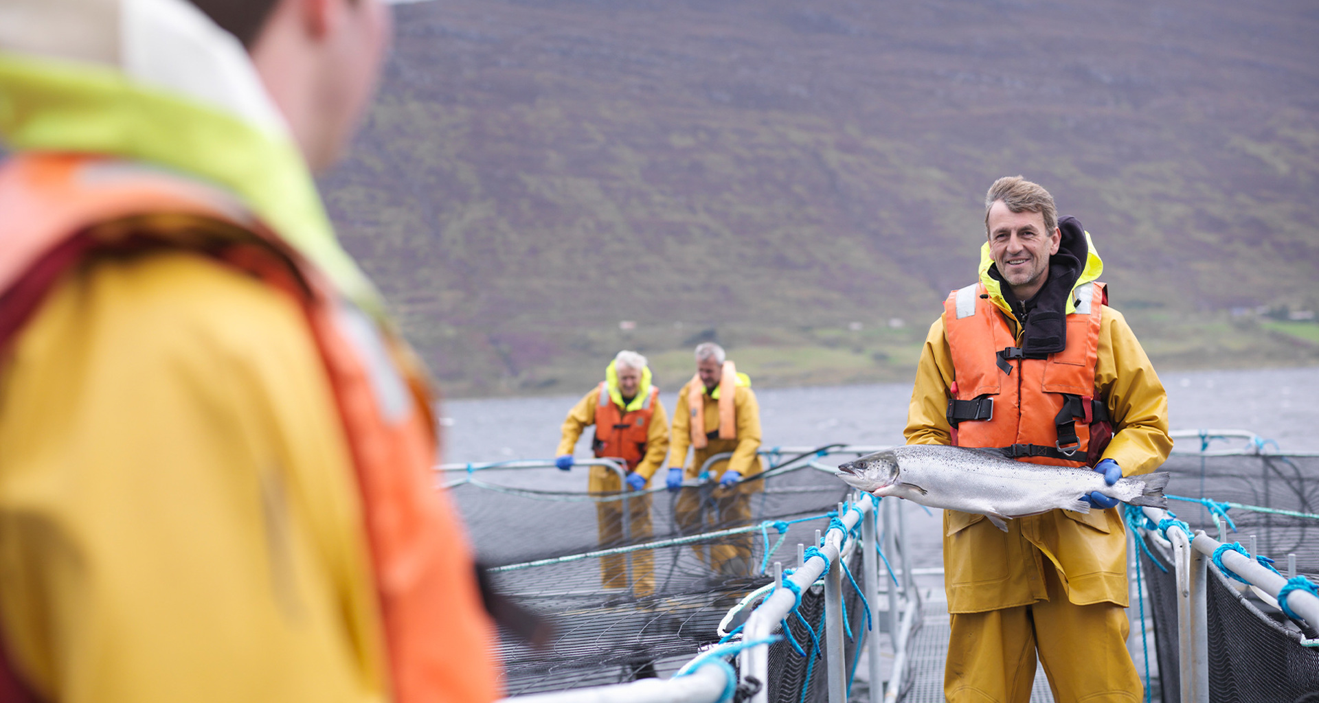 Homme en gilet de sauvetage orange tenant un gros poisson sur une ferme aquacole.
