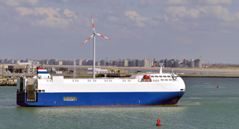 Ship with a wind turbine on deck sailing in a harbor, city skyline behind.