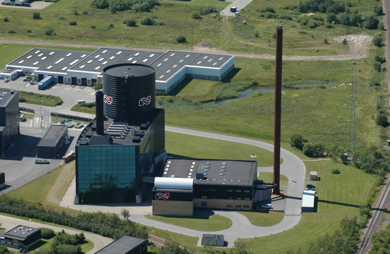 Luftfoto af et industrielt anlæg med en stor cylindrisk struktur og skorsten.