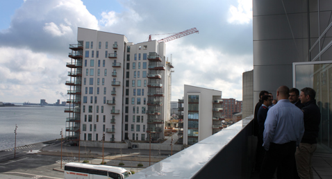 People standing on a balcony overlooking waterfront buildings and a parked bus.