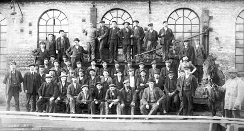 Large group of men in work attire posed outside a brick building.