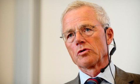 Elderly man in a suit speaking, wearing glasses and a headset microphone.