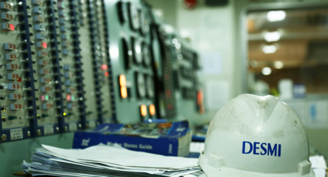 DESMI hard hat on desk in control room with equipment panels.