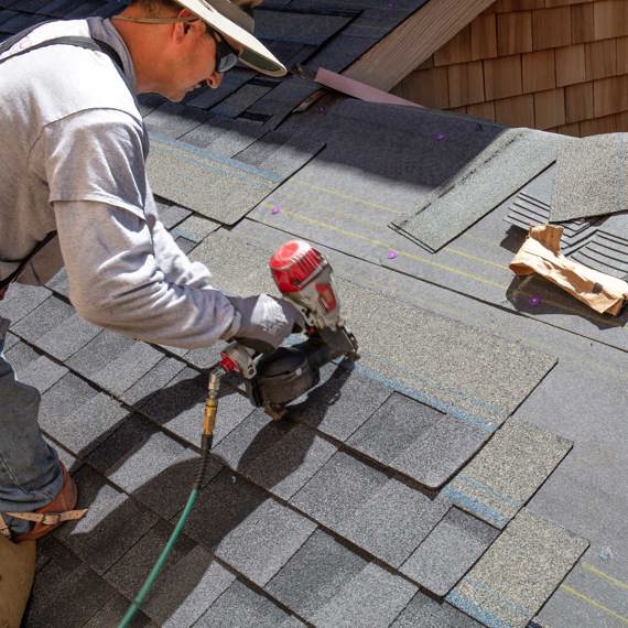 Worker installing asphalt shingles on a roof using a nail gun.