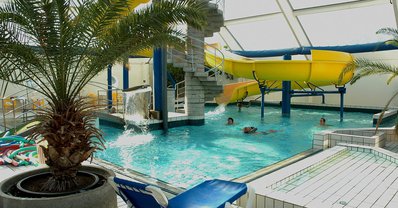 Indoor swimming pool with yellow slide, palm trees, and lounge chairs.