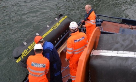Workers in orange suits handle DESMI RO-BOOM equipment on a boat near water.
