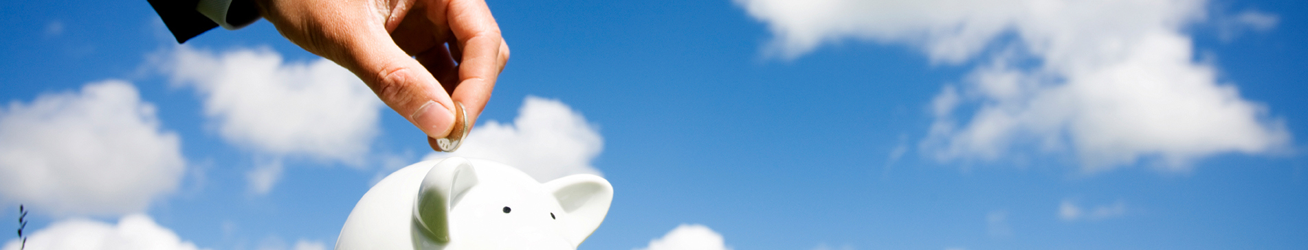 Hand placing coin into white piggy bank against a blue sky background.