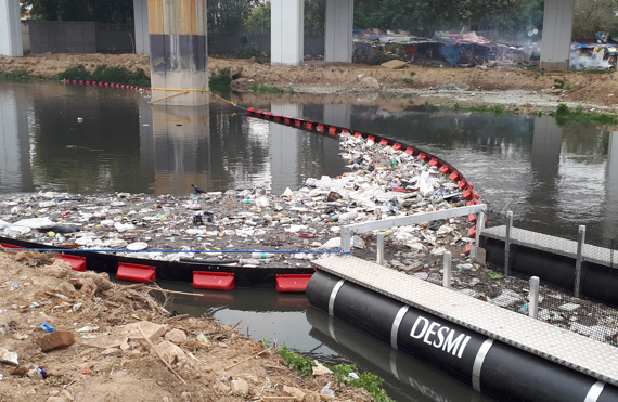DESMI trash barrier containing floating debris in a river under a bridge.