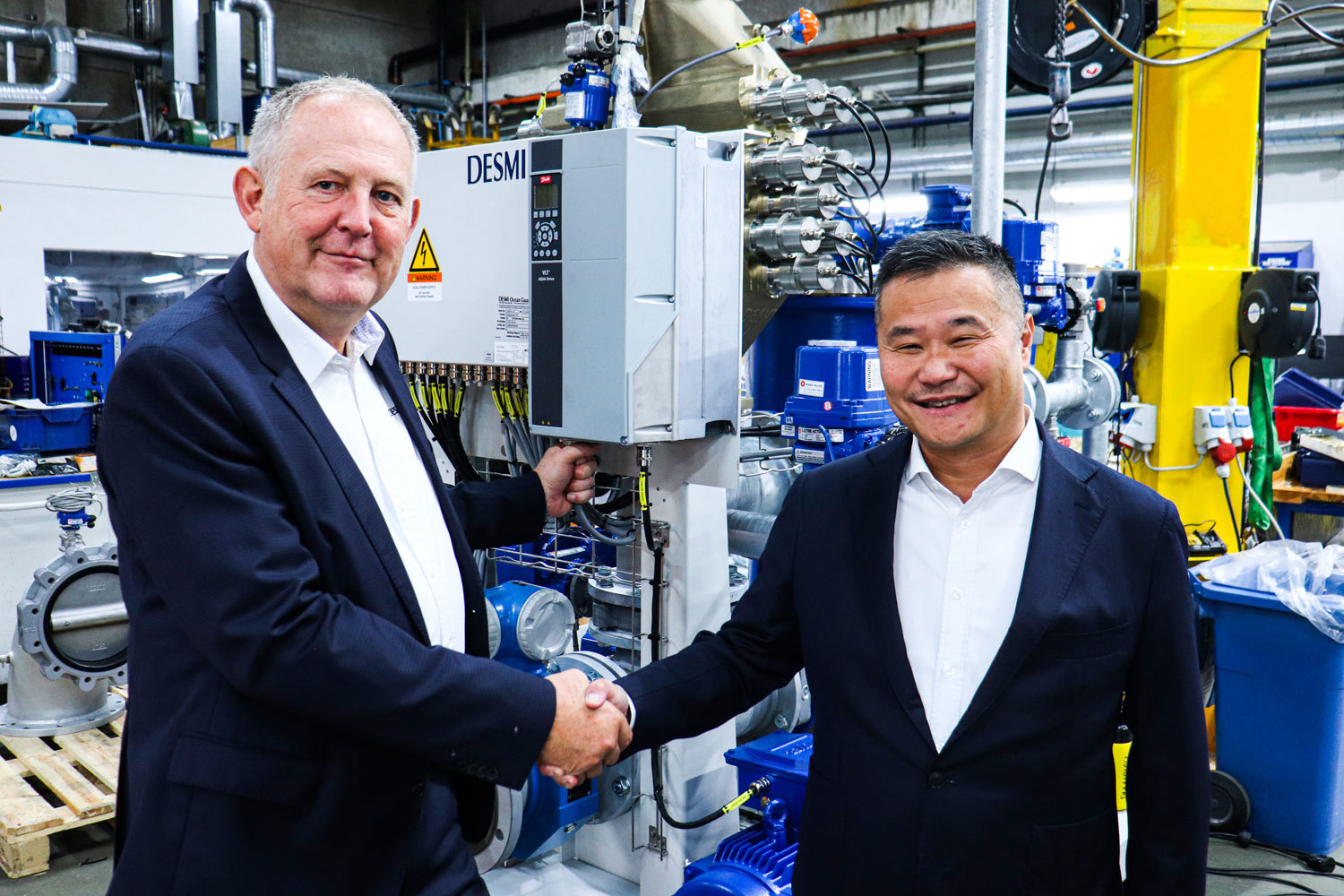 Two men shaking hands in a DESMI facility, showcasing industrial equipment.