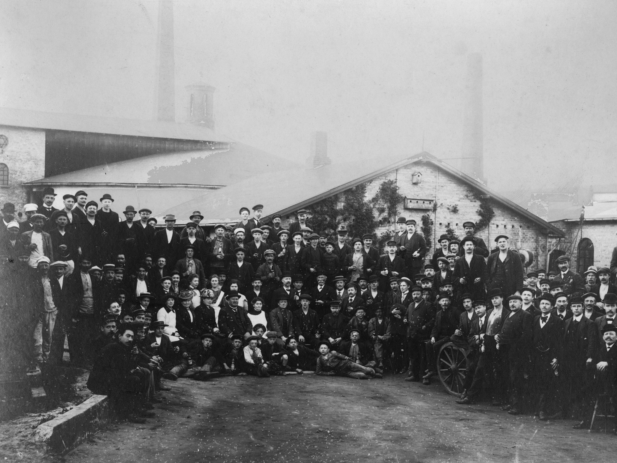 Large group of factory workers posing outside an industrial building in vintage attire.