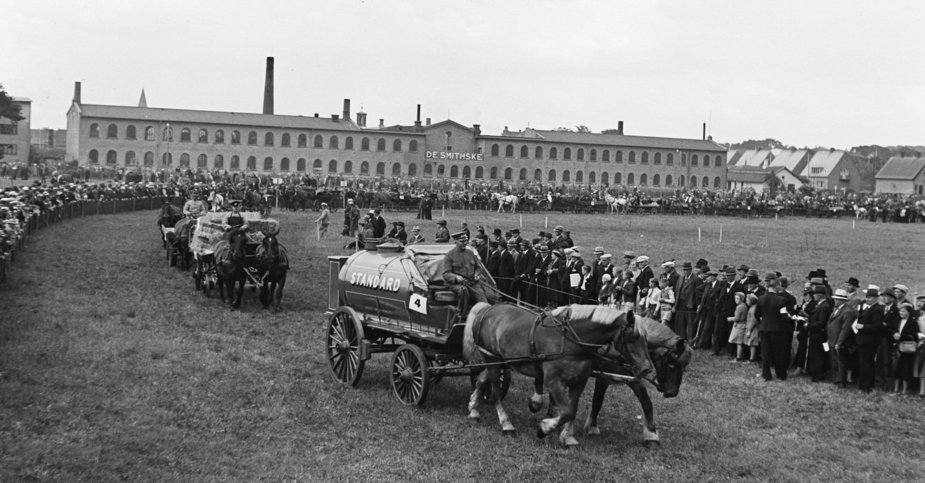 Historic parade with horse-drawn carriages passing by a large crowd and factory building.