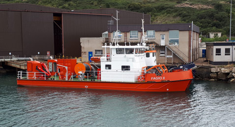 Orange and white vessel "Fagio II" docked near industrial buildings by green hillside.
