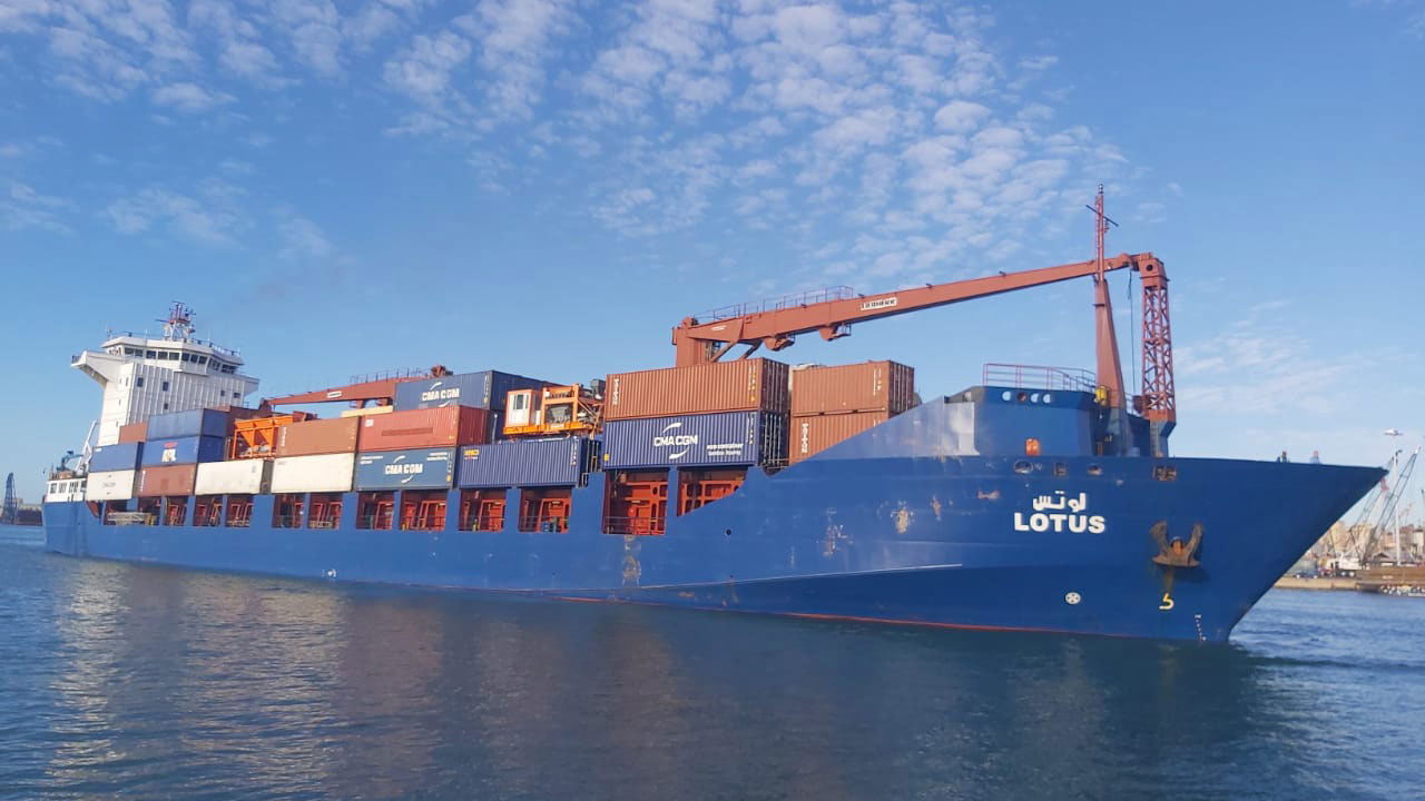 Blue cargo ship "Lotus" carrying stacked containers on a sunny day.