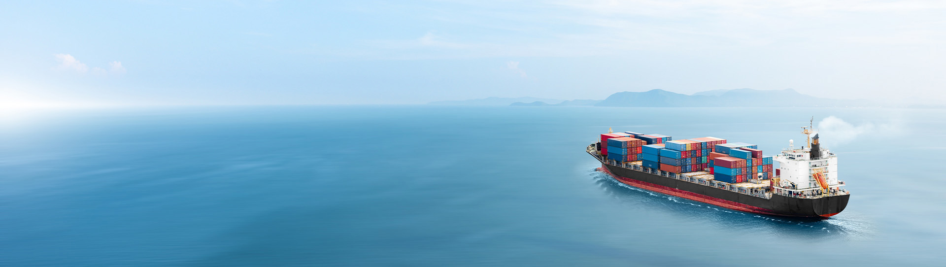Container ship sailing on calm ocean with distant mountains under clear sky.