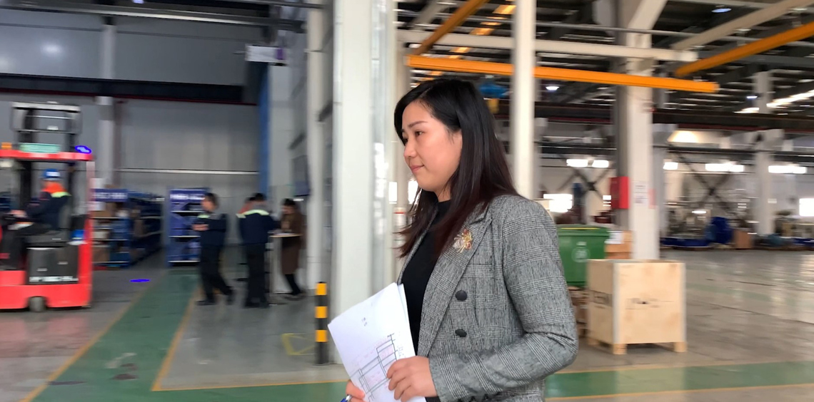 Woman holding documents in an industrial facility with workers and equipment.