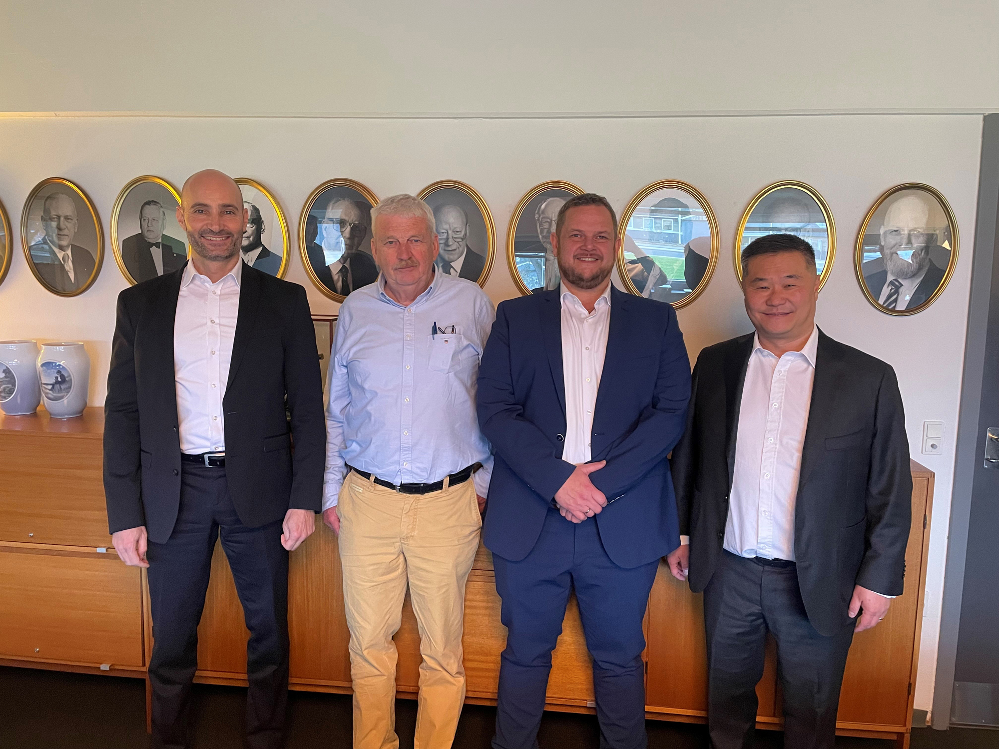 Four men in suits standing in front of framed portraits on a wall.