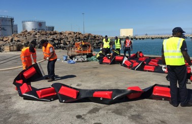 Workers assembling oil containment boom near a dock with storage tanks.