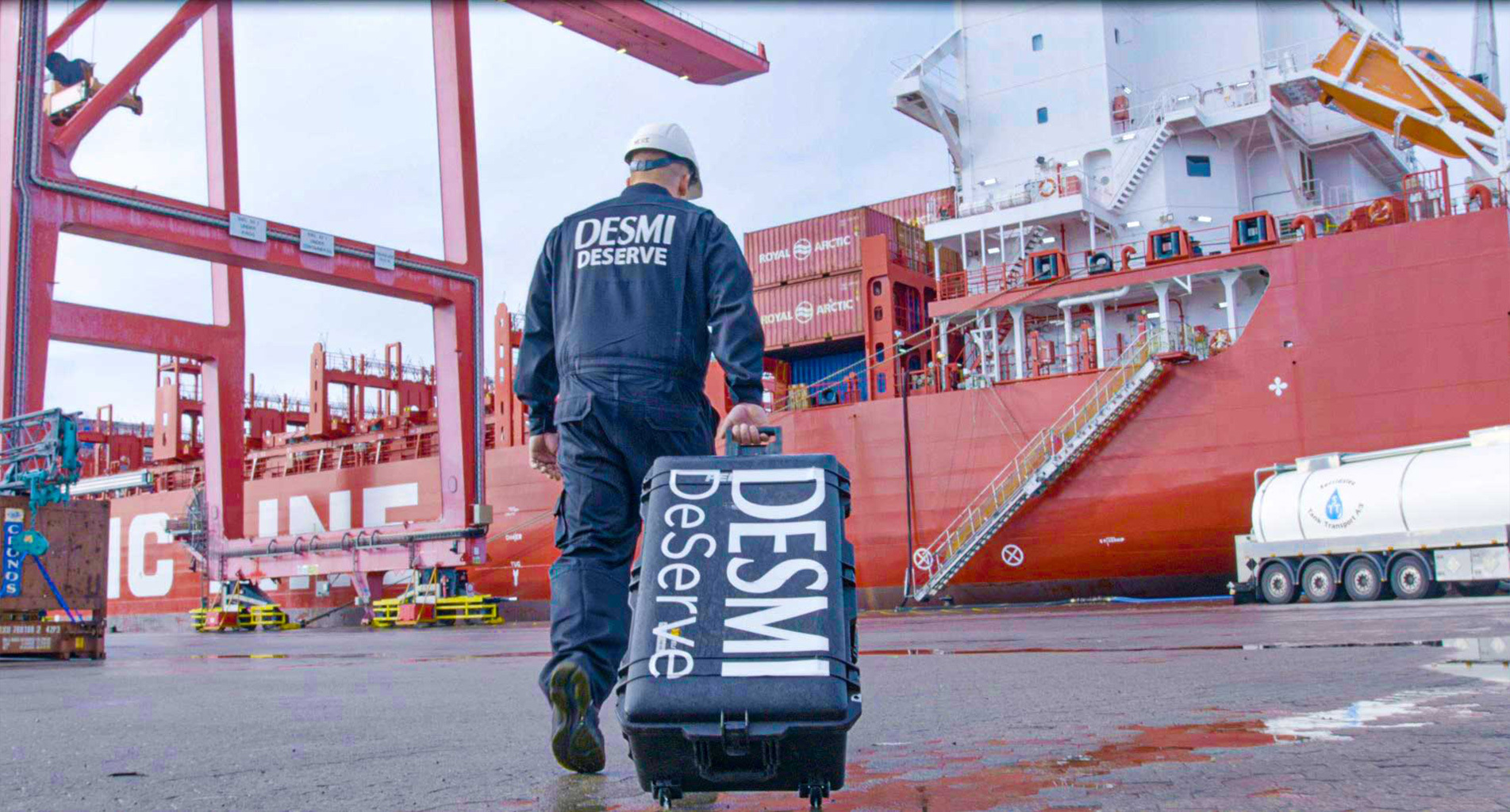 Travailleur en uniforme DESMI tirant une valise près d'un grand cargo.