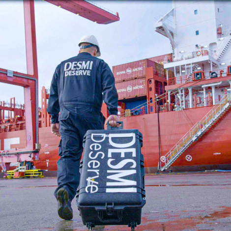 Worker in DESMI uniform pulling a suitcase near a large cargo ship.