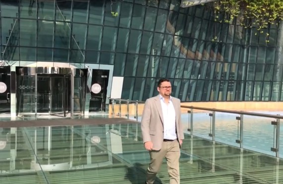 Man walking on a glass bridge outside modern glass building with greenery.