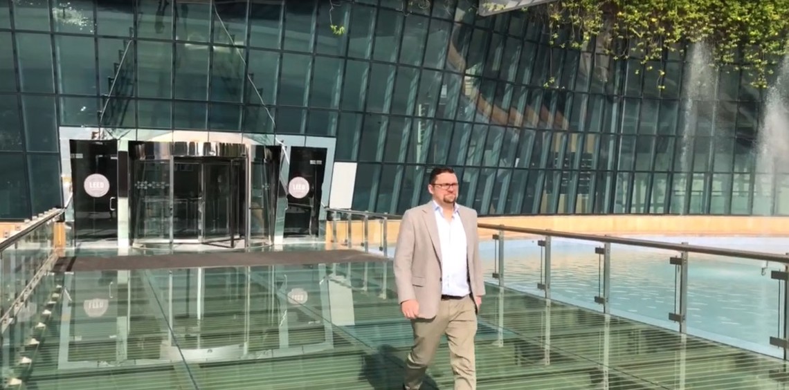 Man walking on a glass bridge outside modern glass building with greenery.
