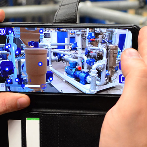 Hands holding a smartphone displaying augmented reality of industrial pipes and machinery.