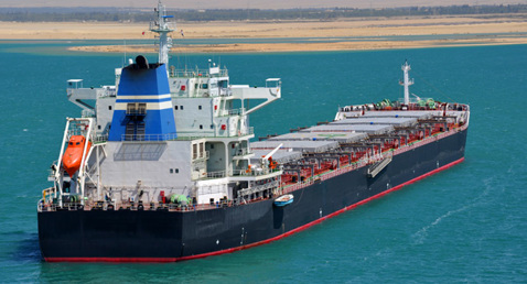 Large cargo ship navigating through calm, turquoise waters near a sandy coastline.